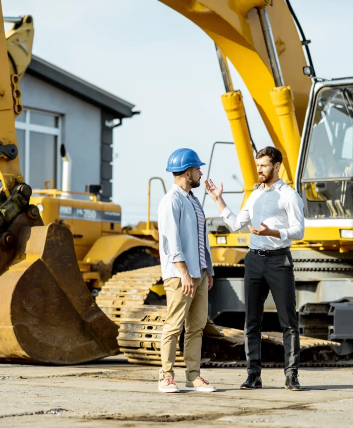 A consultant and a client discussing with an excavator in the background.